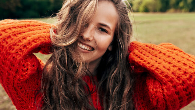 Eine Frau mit langen Haaren und orangefarbenem Strickpulli lächelt