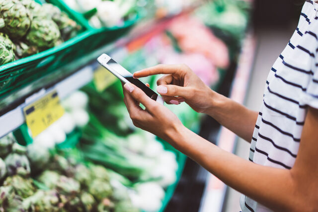 Eine Frau steht vor einem grünen Supermarktregal mit verschiedenen Gemüsesorten und schaut etwas auf ihrem Smartphone nach.