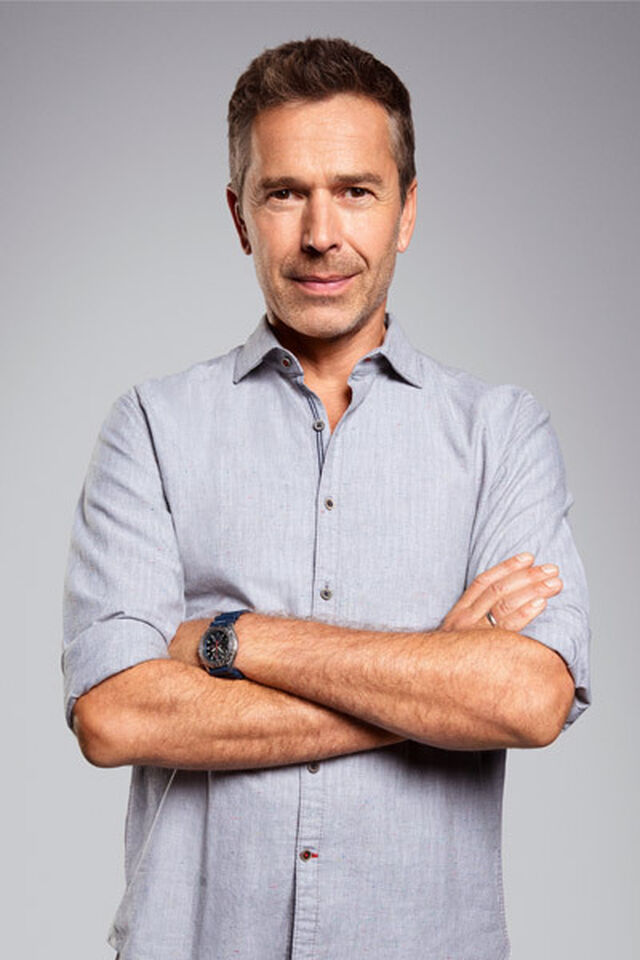Porträt von Dirk Steffens, der in einem hellgrauen, kurzärmeligem Hemd mit verschränkten Armen und einem leichten Lächeln vor einem hellen Hintergrund steht.