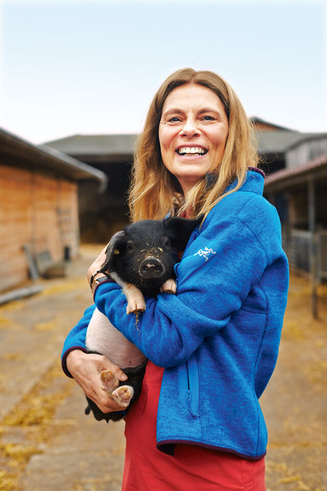 Sarah Wiener trägt eine azurblaue Sportjacke und hält ein kleines schwarz geflecktes Schwein im Arm. Beide gucken in die Kamera. Im Hintergrund sieht man die Umrisse eines Bauernhofs.