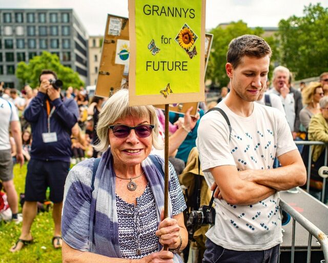 Auf dem Bild ist eine Frau mit grauen Haaren und lila Schal zu sehen, die ein Demoschild mit der Aufschrift Grannys for Future hoch hält.