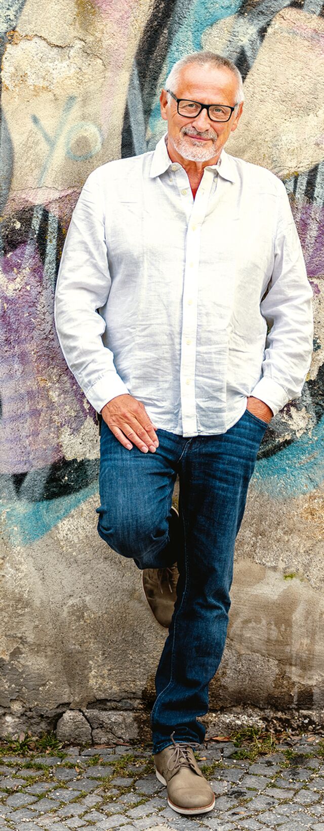 Konstantin Wecker steht in Blue Jeans und weißem Hemd an eine bunte Wand angelehnt. Sein Haar ist weiß, die Brille groß und schwarz.