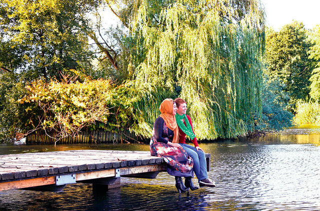 Zwei Frauen sitzen auf einem Holzsteg, der in einen Teich im Park hineinragt. Im Hintergrund Bäume, die am Ufer wachsen