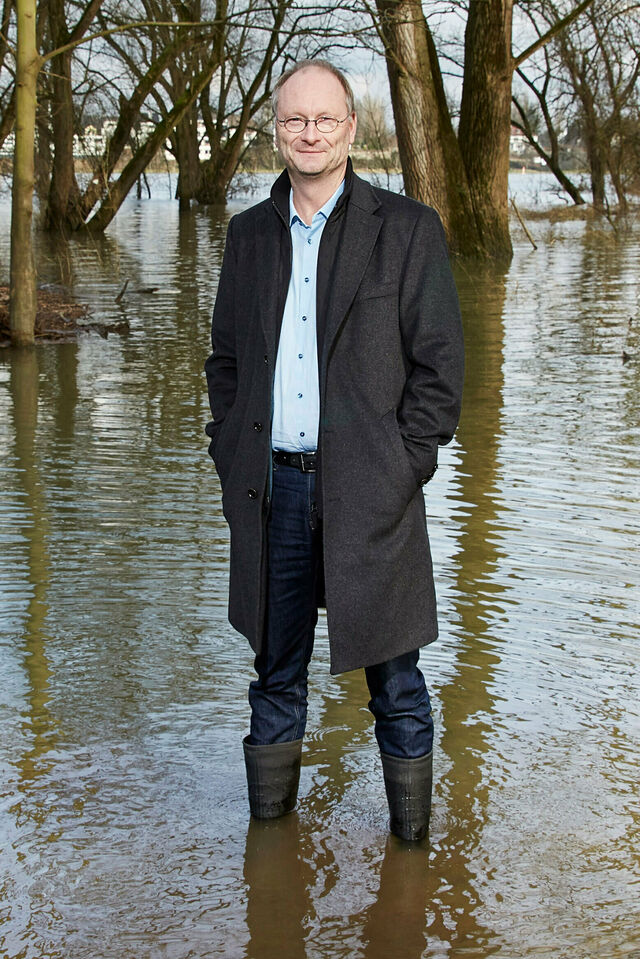 Sven Plöger steht mit Mantel und Stiefeln im Hochwasser vor Bäumen