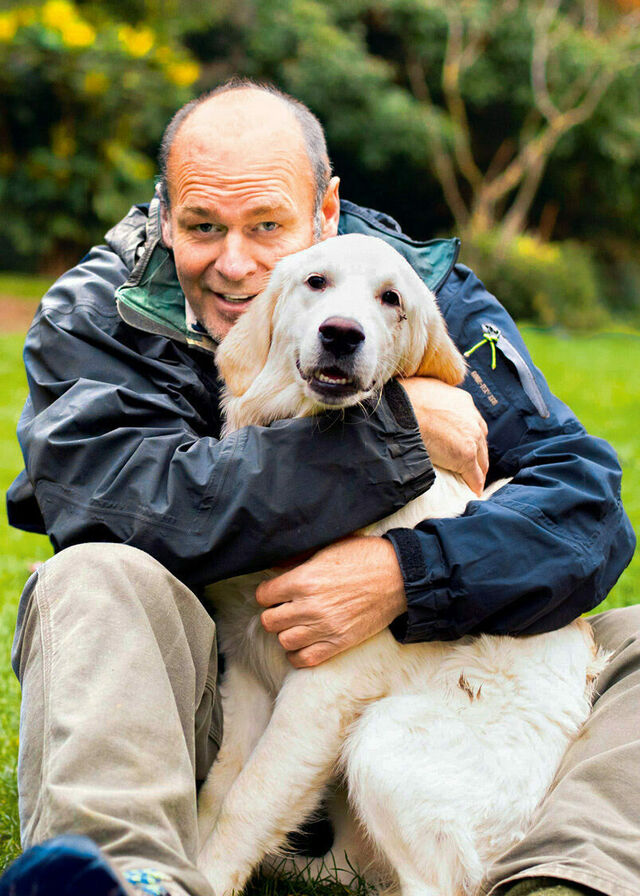 Thomas Henningsen sitzt auf einer Wiese und umarmt seinen Hund