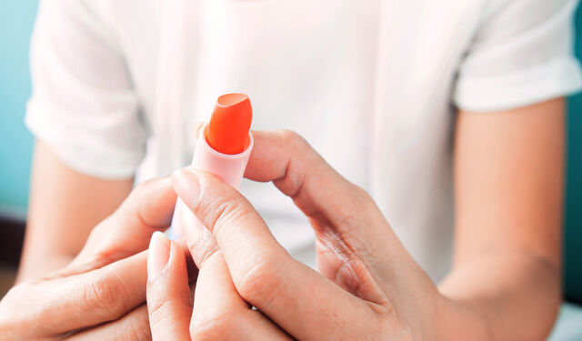 Oberkörper einer Frau, im Vordergrund ihre Hände, in denen sie einen orangefarbenen Lippenstift hält.