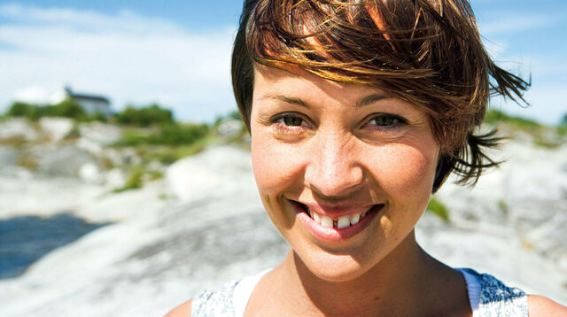 Lächelnde Frau mit Zahnlücke in der Sonne, im Hintergrund Dünen, ein Haus und ein wenig vom Meer.
