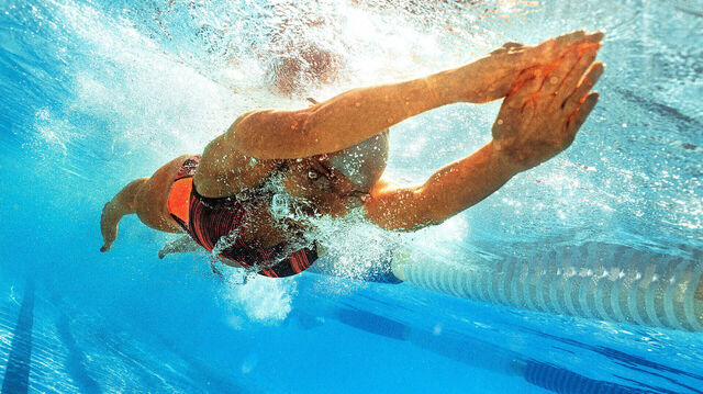 Eine Schwimmerin gleitet durch ein Schwimmbecken. Ihr Körper gleitet durchs Wasser. Sie atmet unter Wasser aus - Luftblasen steigen auf.